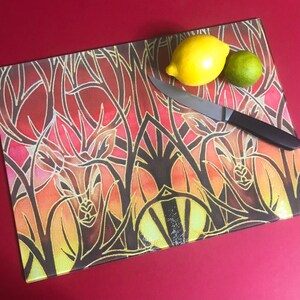 May include: A glass chopping board with a colourful abstract design featuring a stylised floral pattern in shades of red, orange, yellow, and brown. The board is decorated with black lines and a geometric pattern. A knife and two lemons are on top of the board.