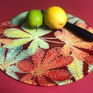 Peut inclure: Une planche à découper ronde avec un fond rouge et un motif de feuilles colorées. Deux citrons, un vert et un jaune, sont sur la planche avec un couteau noir.