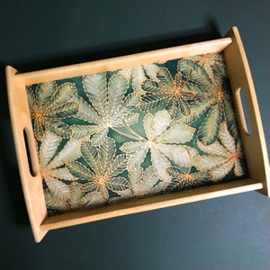 Könnte beinhalten: Ein Serviertablett aus Holz mit einem Blattdesign in Grün und Gold. Das Tablett hat eine rechteckige Form und einen erhöhten Rand.