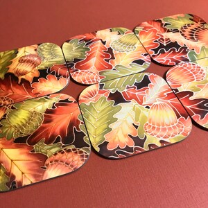 May include: Six square-shaped coasters with an autumnal design. The coasters feature a dark background with a pattern of colourful autumn leaves and acorns.