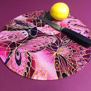 May include: Round glass chopping board with a pink and gold butterfly design. The board is on a purple surface with a knife and a yellow lemon.