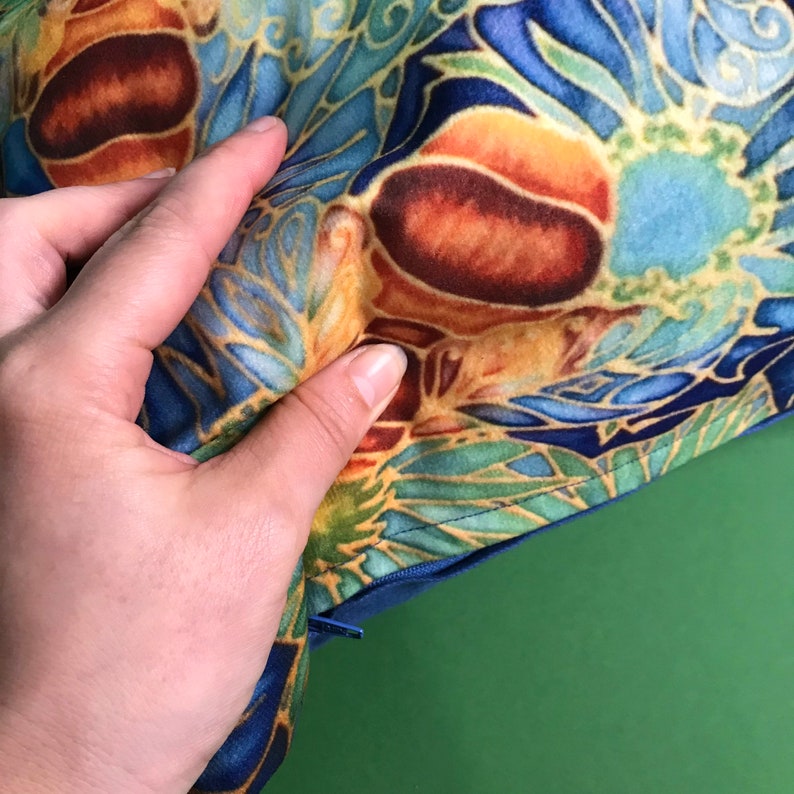 May include: Close-up of a blue and gold fabric with a floral pattern. The fabric has a zipper along the edge and is being held by a hand.