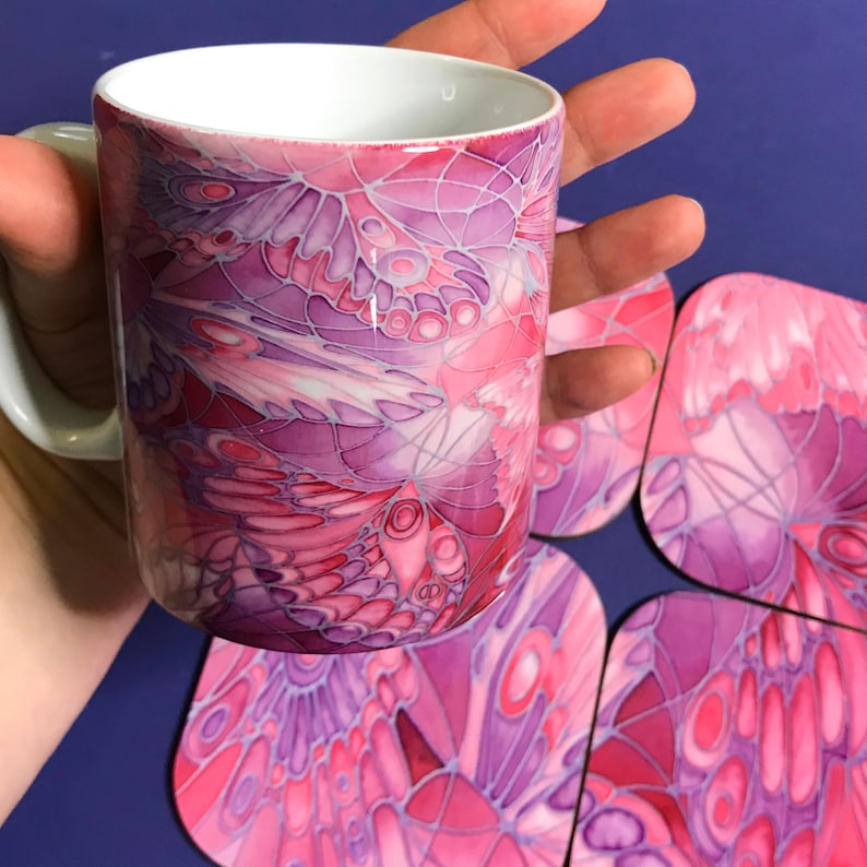 K&ouml;nnte beinhalten: Eine wei&szlig;e Keramiktasse mit einem pinkfarbenen und violetten Schmetterlingsdesign. Die Tasse ist von vier passenden Untersetzern umgeben.