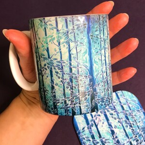 May include: A white ceramic mug with a blue and green forest design. The mug is being held by a hand and a matching coaster is visible in the background.