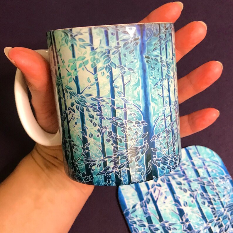 May include: A white ceramic mug with a blue and green forest design. The mug is being held by a hand and a matching coaster is visible in the background.