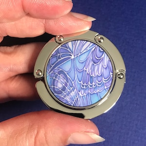 May include: A silver-toned metal purse hook with a round, blue and white patterned design. The pattern resembles a butterfly wing.
