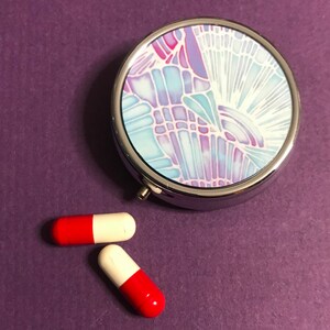 May include: A silver pill box with a colorful abstract design on the lid. Two red and white capsules are next to the pill box on a purple surface.