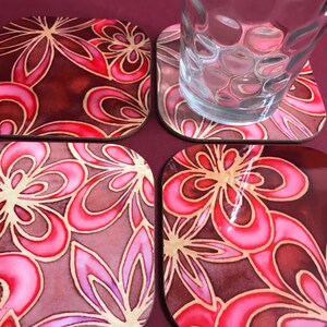 May include: Four square coasters with a floral design. The coasters feature a pink and red floral pattern on a dark brown background.