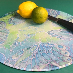 May include: A round glass chopping board with a blue and green butterfly design. A yellow lemon and a green lime are on the board, along with a black handled knife.