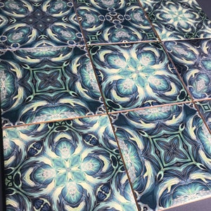 May include: A set of six square ceramic tiles featuring a repeating geometric pattern in shades of blue, green, and white. The tiles have a glossy finish and a subtle gold trim.