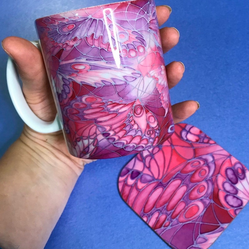 K&ouml;nnte beinhalten: Eine rosa und lila Keramiktasse mit einem Schmetterlingsmuster. Die Tasse wird mit einem passenden Untersetzer geliefert.
