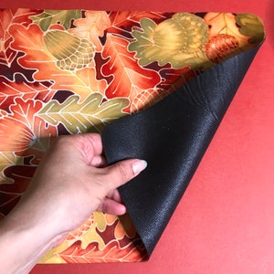 Puede incluir: Un mantel individual con un dise&ntilde;o de hojas de oto&ntilde;o en tonos naranja, amarillo y marr&oacute;n. El mantel individual tiene un respaldo antideslizante negro.