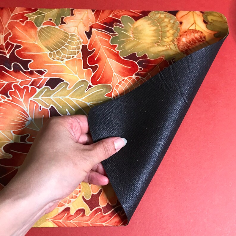 Puede incluir: Un mantel individual con un dise&ntilde;o de hojas de oto&ntilde;o en tonos naranja, amarillo y marr&oacute;n. El mantel individual tiene un respaldo antideslizante negro.