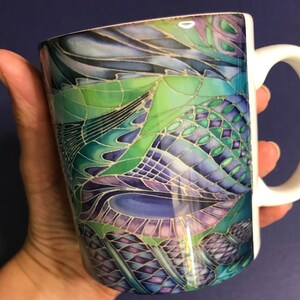 May include: A white ceramic mug with a colourful abstract design featuring shades of green, blue, purple, and gold. The design resembles stained glass or a mosaic.