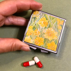May include: A silver pill box with a hinged lid and a floral design featuring yellow daffodils on a green background. Two red and white capsules are on a green surface.