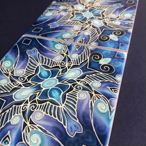 May include: Three square ceramic tiles with a detailed, symmetrical design in shades of blue, teal, and white. The pattern features floral and geometric elements with black outlines. The tiles are arranged side by side, showcasing the repeating artwork.