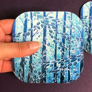 May include: A square blue and white coaster with a design of trees and leaves. The coaster has a rounded edge and is made of a durable material.