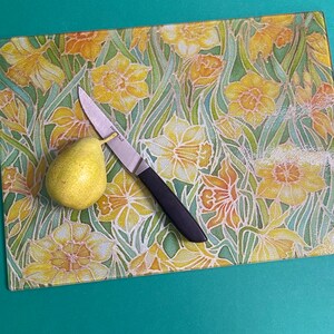 May include: A glass chopping board with a floral pattern featuring yellow daffodils on a green background. A pear and a silver knife are on the board.