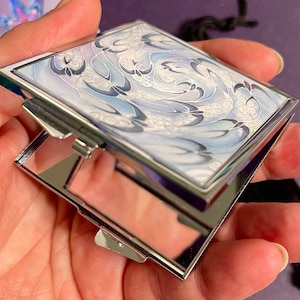 May include: A silver compact mirror with a white and blue swirling design on the lid. The mirror is closed and the lid is facing the camera.