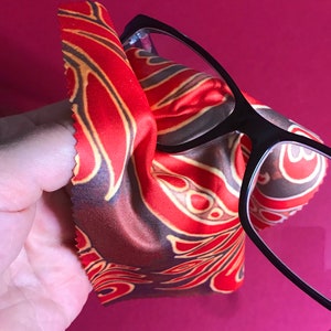 May include: A close-up of a pair of black-framed glasses being cleaned with a red and gold patterned microfiber cloth.