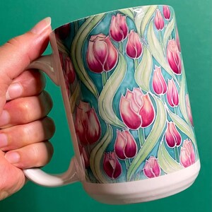 Può includere: Tazza in ceramica bianca con un design floreale con tulipani rosa e foglie verdi su uno sfondo blu chiaro.