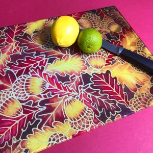 May include: A glass chopping board with an autumnal design featuring red, yellow and gold leaves and acorns. A lemon and a lime are on the board, along with a black-handled knife.