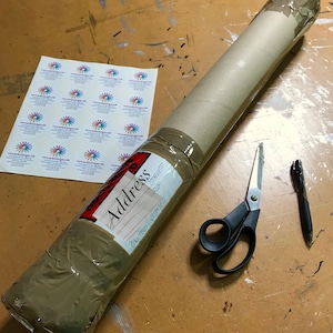 May include: A brown cardboard tube wrapped in brown packing tape with a red label that says "Address" and an address written on it. The tube is sitting on a wooden surface with a pair of black scissors and a black pen.
