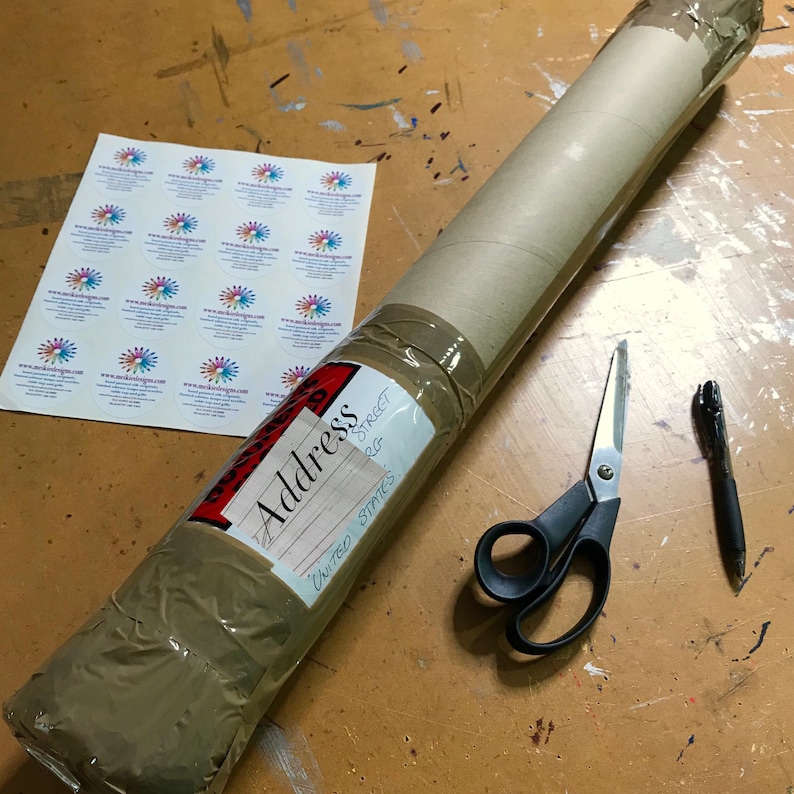 May include: A brown cardboard tube wrapped in brown packing tape with a red label that says "Address" and an address written on it. The tube is sitting on a wooden surface with a pair of black scissors and a black pen.