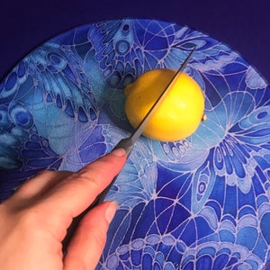 May include: A yellow lemon is being cut in half with a silver knife on a blue and white glass chopping board with a butterfly design.