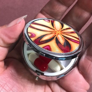 May include: A silver pill box with a colourful floral design on the lid. The box is open and contains two pills.