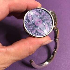 May include: A silver-toned metal purse hook with a round, hinged, decorative top featuring a purple and white butterfly design.