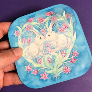 May include: Two white rabbits with pink eyes are nestled together inside a heart-shaped wreath of green leaves and pink flowers on a blue background.