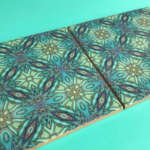 May include: Two square ceramic tiles featuring an intricate geometric pattern in shades of blue, green, and gold. The pattern is symmetrical and resembles a kaleidoscope.