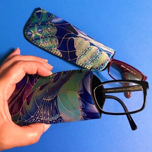 Op de afbeelding: Twee gepatroneerde brillenetuis van stof, een blauw en groen met een bloemenpatroon en een paars en groen met een geometrisch patroon. Een paar zwarte brillen met een bordeauxrode montuur is gedeeltelijk zichtbaar.