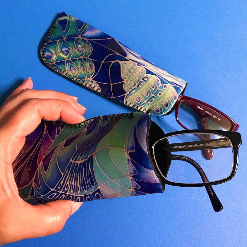 Op de afbeelding: Twee gepatroneerde brillenetuis van stof, een blauw en groen met een bloemenpatroon en een paars en groen met een geometrisch patroon. Een paar zwarte brillen met een bordeauxrode montuur is gedeeltelijk zichtbaar.