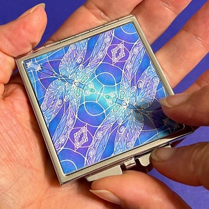 Può includere: Uno specchio compatto argentato con un design di libellula blu e viola. Lo specchio ha un coperchio incernierato e una piccola maniglia decorativa.