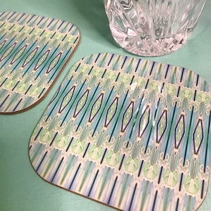 May include: Two square coasters featuring a geometric design in shades of blue, green, and white. The coasters have a light brown wooden base.