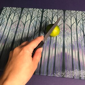 May include: A hand holding a silver knife cutting a green lime on a glass chopping board with a purple and white forest design.