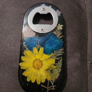 Puede incluir: Abridor de botellas negro con un abridor de metal plateado. El abridor está decorado con una margarita amarilla y flores azules, junto con vegetación seca, encapsulada en resina.