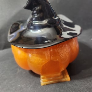 May include: Orange pumpkin-shaped container with a black witch hat lid. The container has a glittery finish and a spiderweb design.