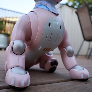 May include: A pink robotic dog with silver joints and a blue and white patterned bandana. The dog is sitting on a wooden deck.
