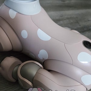 May include: A close-up of a pink robotic dog's leg and body, with white polka dots. The dog has a white collar and a camera eye. The logo "Virtual Paws" is visible in the bottom right corner.