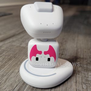 May include: A white robot with a pink face and two black eyes. The robot is on a white base with a gray ring around the edge. The robot has a white head with a gray button on top. The text "@VIRTUAL_PAWS" is visible on the base.