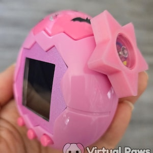 以下が含まれることがあります： ピンク色の卵型の電子ペットゲーム。星型のアクセサリーが付いています。小さな画面とピンク色のボタンがあります。デバイスは手で持たれています。