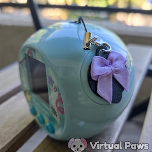 May include: A light blue handheld electronic pet game with a screen and buttons. A purple bow is attached to the side. The device has a black strap and silver-colored metal clasp. The screen displays colorful graphics.