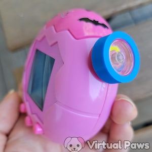 May include: A pink, egg-shaped toy with a blue and clear lens. The toy has a screen and small pink feet. The words "Virtual Paws" are printed on the bottom right of the image.