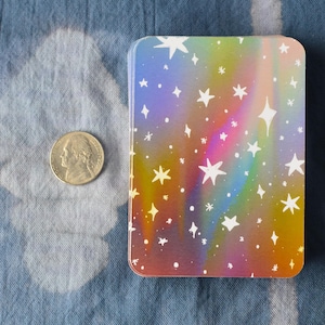 Pode incluir: Um baralho retangular com um fundo de gradiente arco-&iacute;ris hologr&aacute;fico e um padr&atilde;o de estrelas brancas de v&aacute;rios tamanhos. Uma moeda americana de cinco centavos est&aacute; colocada ao lado para escala, medindo 2,12 cm de di&acirc;metro.
