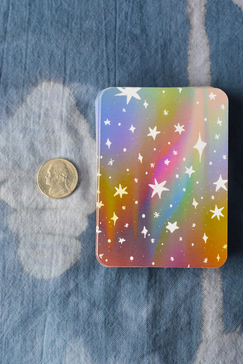 Pode incluir: Um baralho retangular com um fundo de gradiente arco-&iacute;ris hologr&aacute;fico e um padr&atilde;o de estrelas brancas de v&aacute;rios tamanhos. Uma moeda americana de cinco centavos est&aacute; colocada ao lado para escala, medindo 2,12 cm de di&acirc;metro.