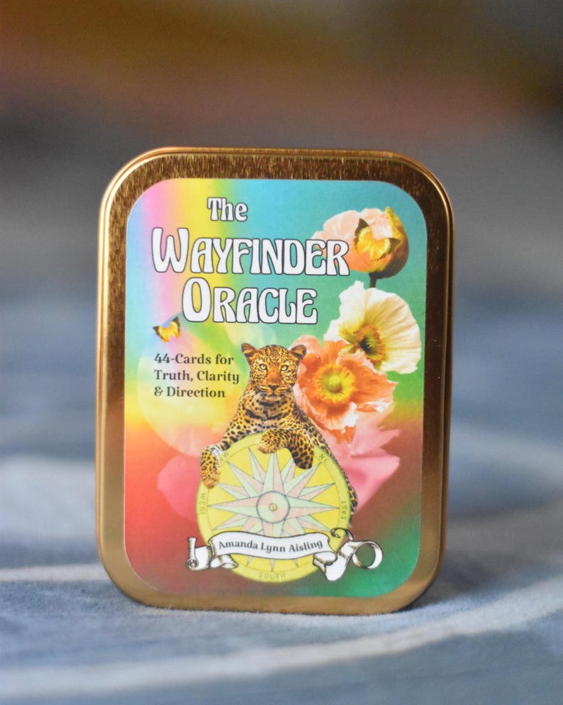 Pode incluir: Uma lata retangular de metal na cor dourada contendo um baralho intitulado The Wayfinder Oracle. O r&oacute;tulo apresenta um leopardo descansando sobre uma b&uacute;ssola, cercado por flores e borboletas coloridas, com o texto 44-Cards for Truth, Clarity & Direction e o nome da autora Amanda Lynn Aisling.