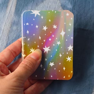 Pode incluir: Um baralho de cartas retangulares com um fundo hologr&aacute;fico de cores do arco-&iacute;ris e um padr&atilde;o repetitivo de estrelas brancas desenhadas &agrave; m&atilde;o de v&aacute;rios tamanhos espalhadas pela superf&iacute;cie de cada carta no monte.